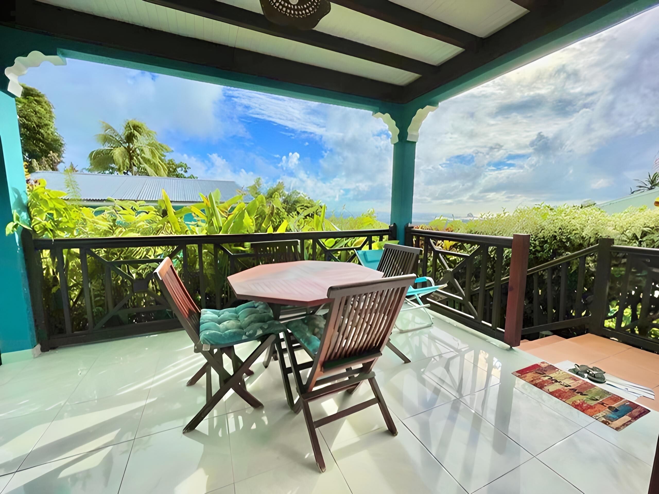 A covered outdoor area with a table and chairs, a tiled floor, and a view of greenery and a cloudy sky.