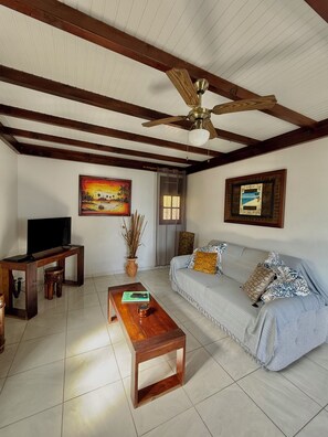 A living room with a wooden ceiling, a ceiling fan, a television on a wooden stand, a sofa with patterned cushions, and a coffee table with a book and a glass. - LES VILLAS PAMEYAQUE/ LA VILLA PAME with a pool and a view of the Caribbean Sea (Rivière-Salée)