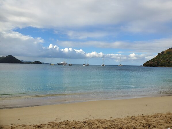 A beach with clear water, a sandy shore, and several sailboats anchored in the distance.