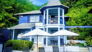 A two-story house with a balcony, outdoor seating area with chairs and umbrellas, and a garden. - Panoramic Views as Sunsets await, Close to Beaches, Sandals Golf and Cabot Golf (Cap Estates)