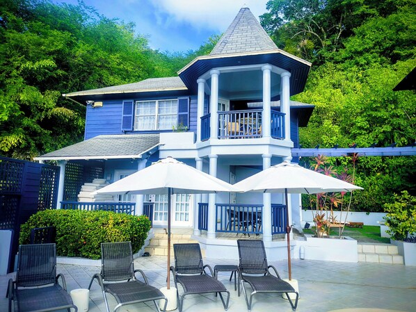 A two-story house with a balcony, outdoor seating area with chairs and umbrellas, and a garden. - Panoramic Views as Sunsets await, Close to Beaches, Sandals Golf and Cabot Golf (Cap Estates)