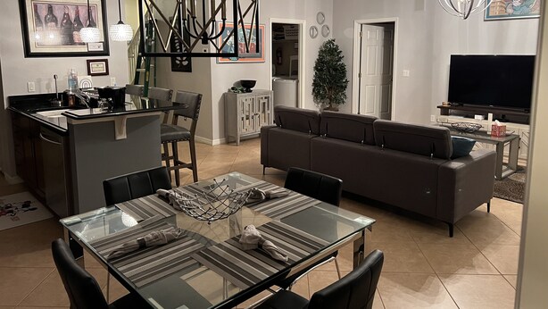 A modern living room with a glass dining table, a grey sofa, and a kitchen area with a sink and stove.