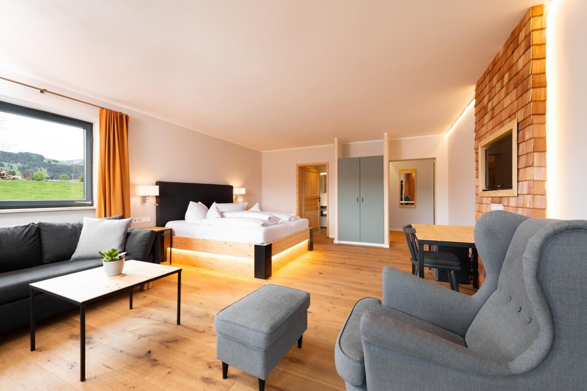 Quarto de hotel moderno com cama grande, sofá, mesa de jantar e vista para o exterior.