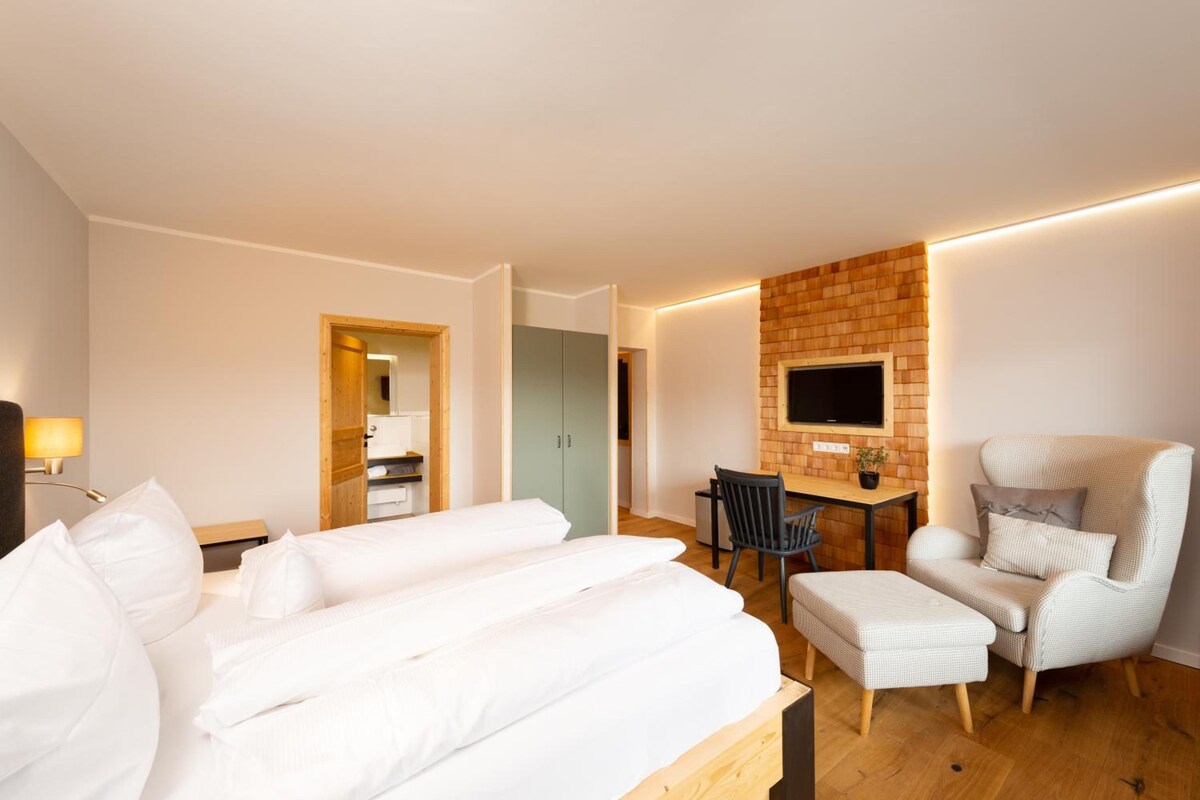 Quarto de hotel com uma cama, uma escrivaninha, uma cadeira, uma televisão presa a uma parede de tijolos e um abajur.
