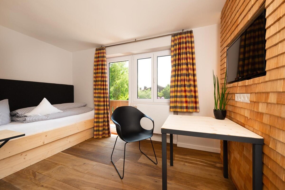 Quarto com cama, mesa, cadeira, janela com cortinas e uma planta.