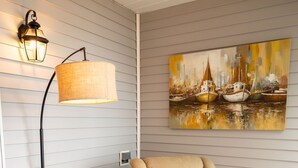 A room with a beige armchair, a glass-top table, a floor lamp, and a painting of boats on the wall.