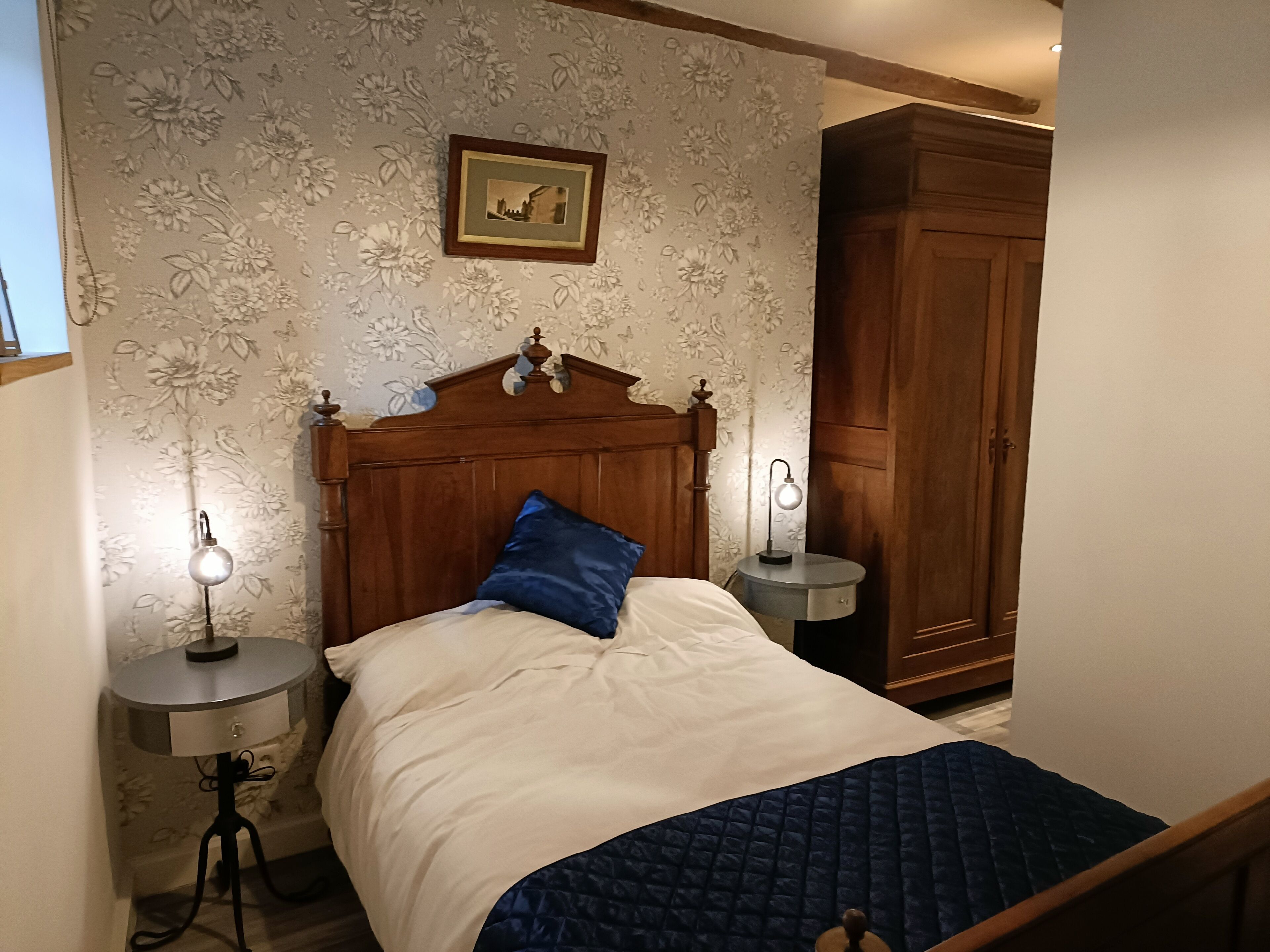 A bedroom with a wooden headboard, a bed with a blue pillow, two bedside tables with lamps, and a wardrobe.