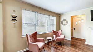 Une pièce avec deux fauteuils roses, une petite table ronde et une grande fenêtre avec des stores.
