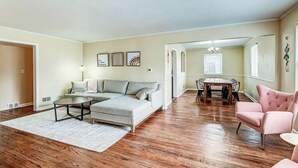 Un vaste salon comprenant un canapé d’angle, une table basse ronde et un fauteuil rose. On y trouve également un coin repas avec une table et des chaises en bois. Le sol est recouvert de parquet et les murs sont peints dans des tons clairs.