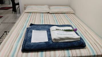 Ein ordentlich bezogenes Bett mit gestreifter Bettwäsche, ein gefaltetes Handtuch, eine Willkommenskarte und eine kleine violette Blume.