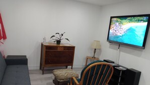 Uma sala de estar com um sofá cinza, uma cadeira de madeira, uma pequena mesa de madeira com uma luminária, uma TV na parede e um armário de madeira com uma planta.