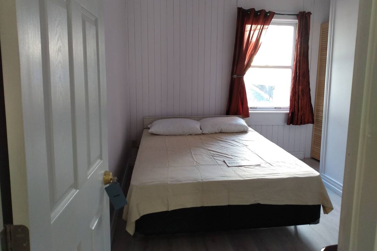 Ein Schlafzimmer mit einem Bett, weißen Wänden, einem Fenster mit Vorhängen und einer Tür.