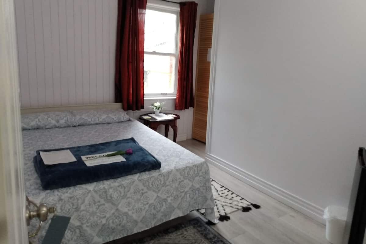Ein Schlafzimmer mit einem Bett, einem Nachttisch, einem Fenster mit Vorhängen und einem kleinen Tisch mit Blumen in einer Vase.
