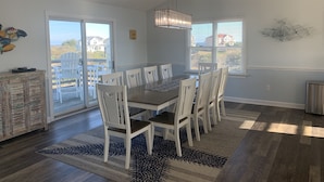 A dining area with a large table, white chairs, a chandelier, and a view of the outside. - Water Music OBX:  Ocean views, easy beach access, private pool.  Pet friendly. (Corolla)