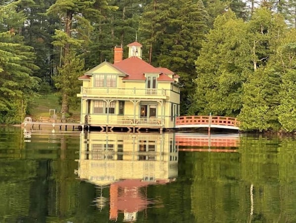 A large, multi-story house with a red roof and a balcony, surrounded by dense trees and a calm lake.