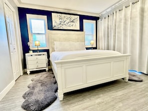 Une chambre avec un lit blanc, deux tables de chevet avec des lampes, une grande fenĂȘtre avec des stores, un tableau au mur et un tapis en fourrure sur le sol.