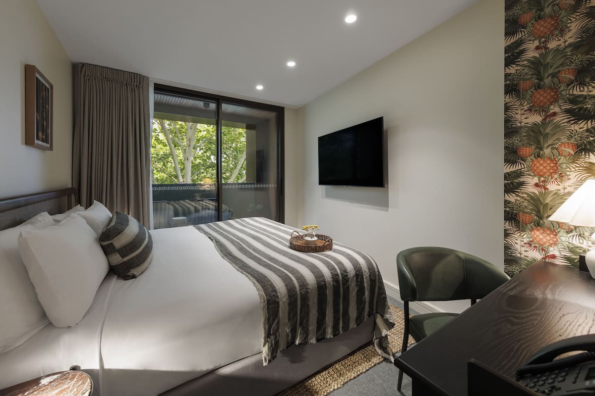 A hotel room with a large bed, a desk, a chair, a television, and a window with a view of trees.