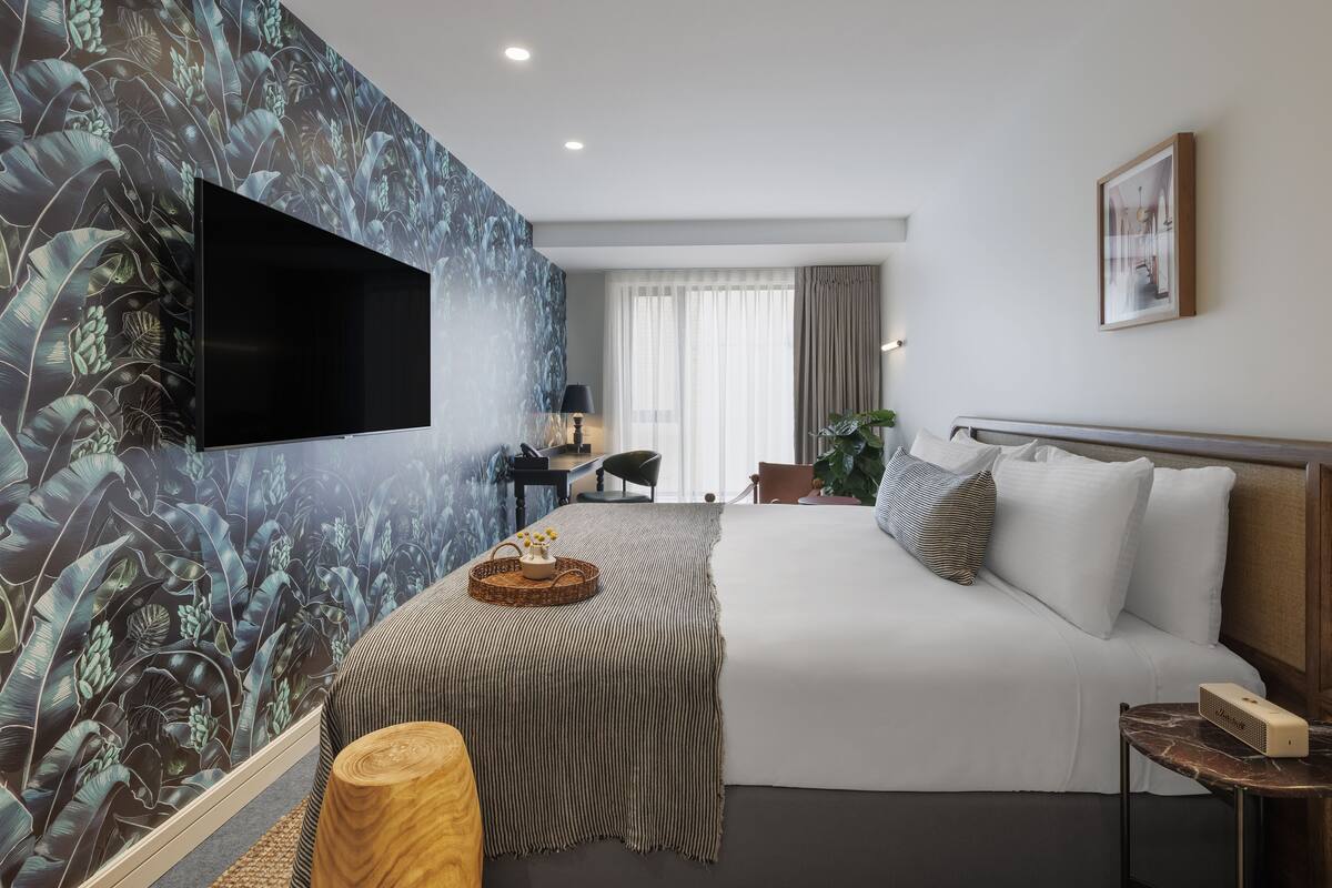 A modern hotel room with a large bed, a flat-screen TV mounted on the wall, a desk with a chair, and a large window with curtains.
