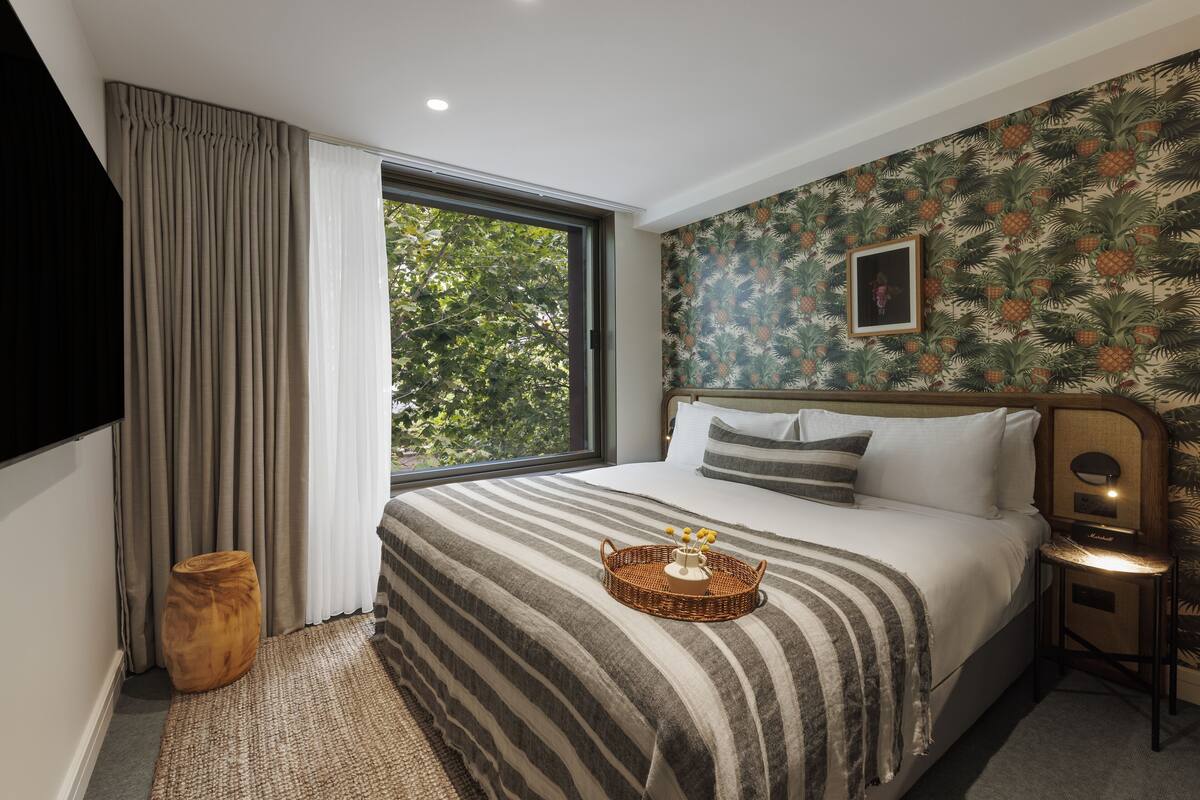 A hotel room with a large bed, a window with curtains, a wall with a tropical wallpaper, and a small table with a lamp.