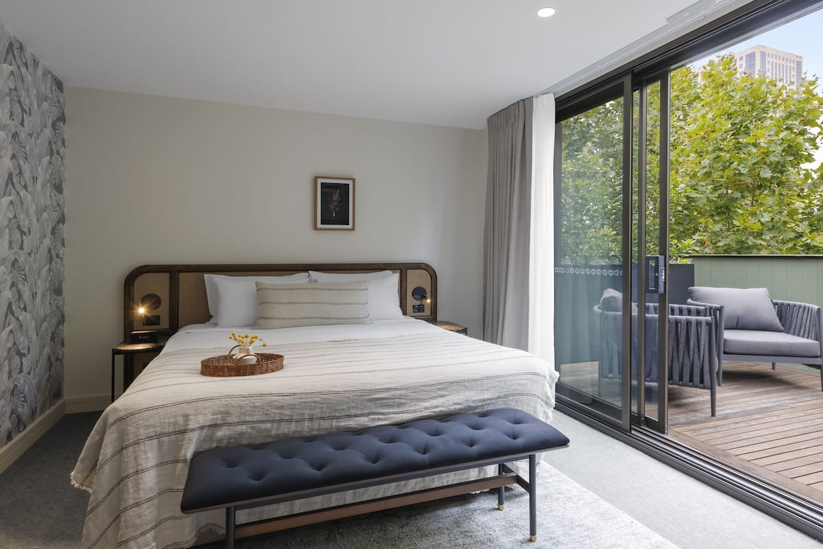 A bedroom with a large bed, a bench, a nightstand, and a view of a balcony with outdoor furniture.