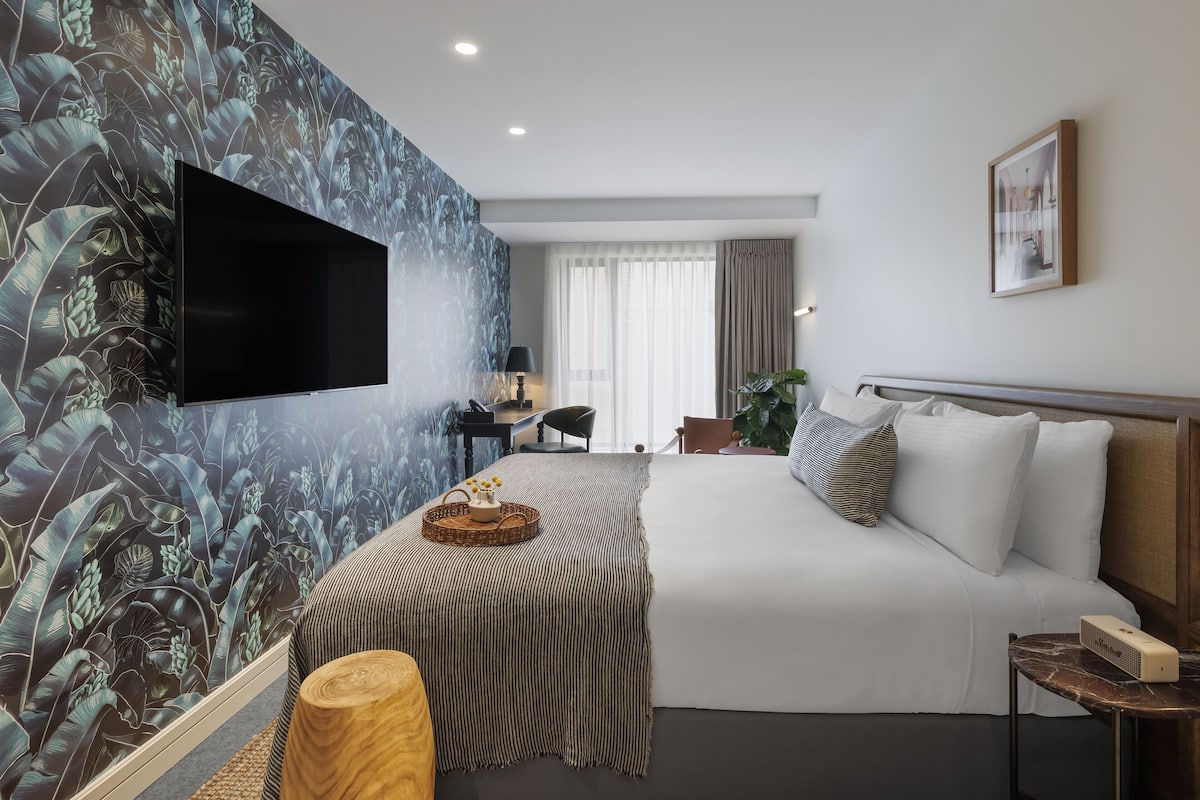 A hotel room with a large bed, a flat-screen TV mounted on the wall, a desk with a chair, and a window with curtains.