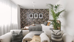 Un dormitorio con una cama, dos sillones, una mesita de centro, una planta grande en maceta y una pared decorada con cuadros.