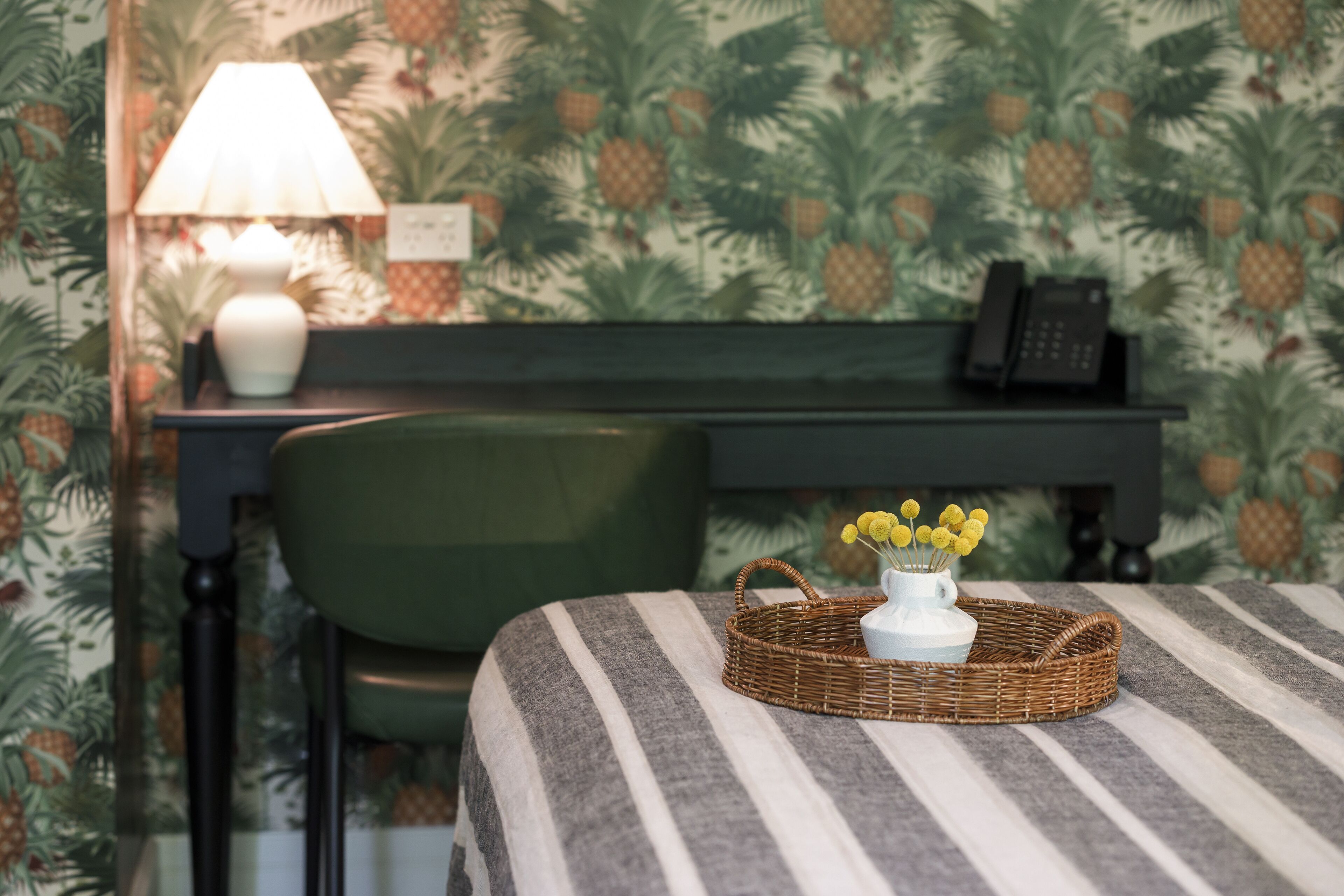 Una habitación con una colcha a rayas, una cesta de mimbre con flores amarillas, un jarrón blanco, una lámpara, un teléfono y una silla.