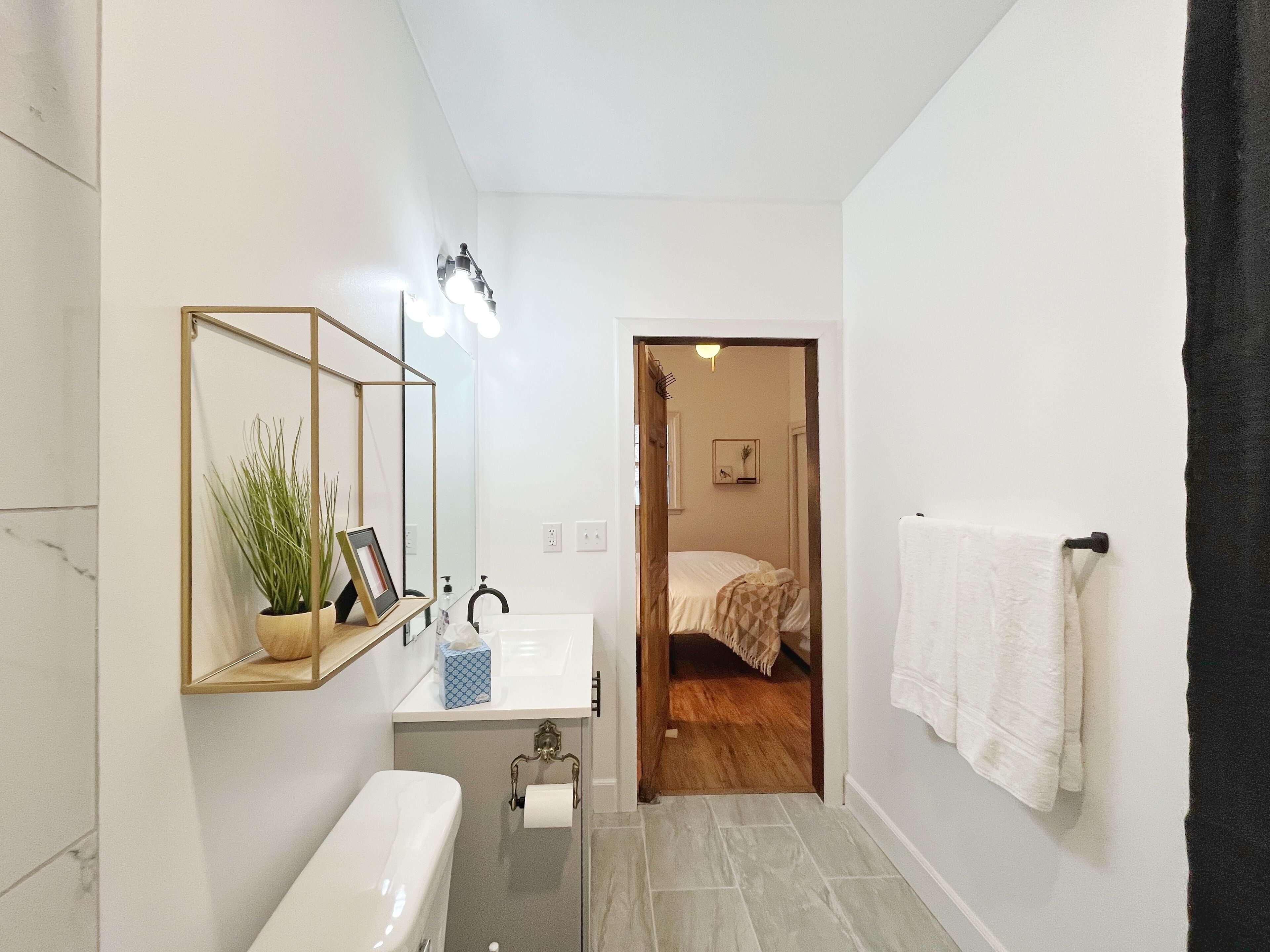 Une salle de bain moderne avec un lavabo blanc, un miroir et des toilettes. Une porte donne sur une chambre à coucher où se trouvent un lit et un tableau accroché au mur.