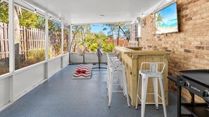 Un patio techado al aire libre con zona de bar, taburetes de metal blanco, una parrilla y vistas al jardín trasero.