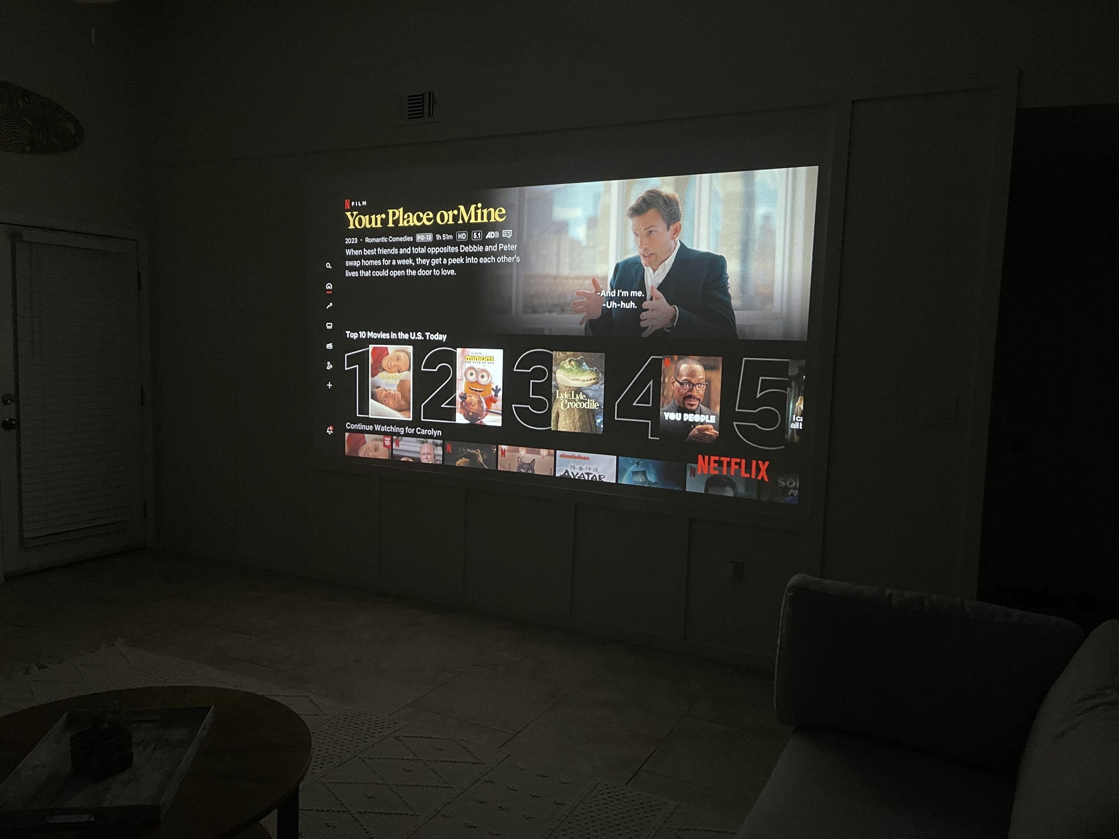 Una pantalla de proyector mostrando la película 'Your Place or Mine' con una lista de las películas más populares en la plataforma Netflix.