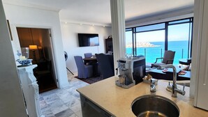 Une chambre d’hôtel moderne dotée d’un coin cuisine, d’une table à manger avec des chaises, d’un téléviseur à écran plat fixé au mur et d’une grande fenêtre offrant une vue sur la mer.