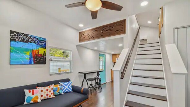 Un salon moderne avec un canapé, des coussins colorés, un ventilateur de plafond et un escalier.