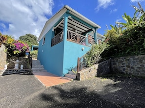 Um edifício de dois andares com fachada azul, escadaria de pedra e um pequeno jardim com flores cor-de-rosa.