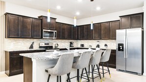 A modern kitchen with dark wood cabinets, stainless steel appliances, a granite countertop, and bar stools. - 10BR Villa w/ Pool, Theater Loft & Cabanas (Davenport)