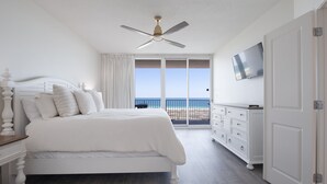 A bedroom with a bed, a ceiling fan, a TV, a dresser, and a view of the beach.