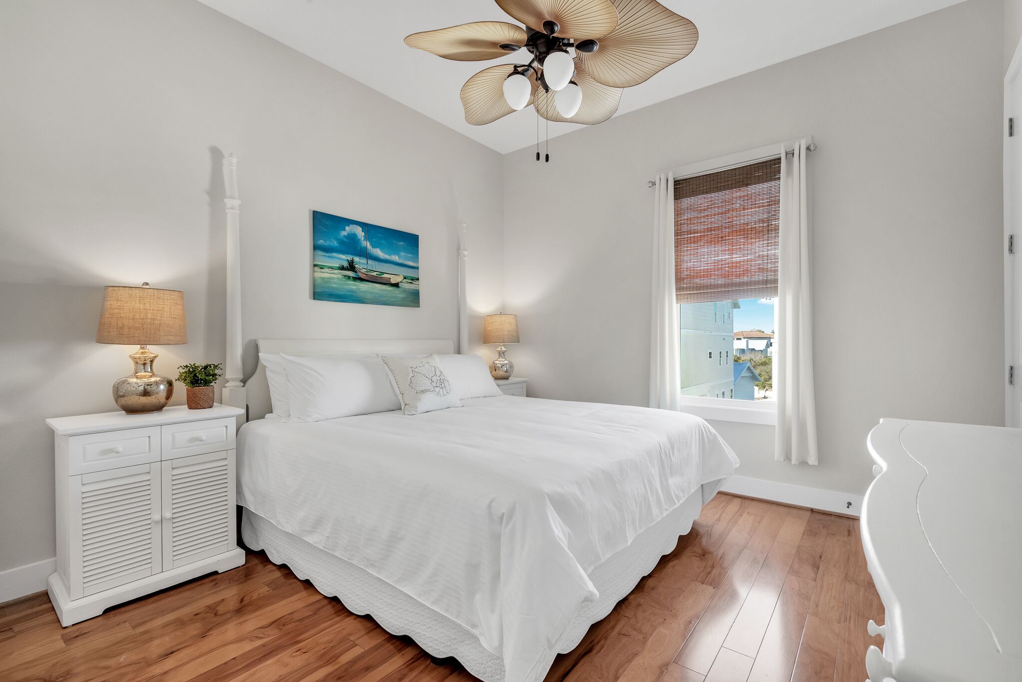 Quarto com cama, mesas de cabeceira, ventilador de teto, janela com persianas e um quadro na parede.