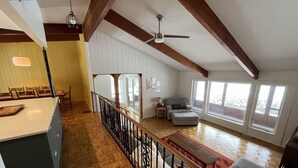 Ein geräumiges Wohnzimmer mit hoher Decke, Holzbalken, einer Essecke mit Stühlen, einem Sofa und einem großen Fenster, das für natürliches Licht sorgt.