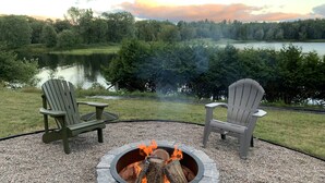 Ein Steinkamin mit zwei Adirondack-Stühlen vor einem See und Wald.