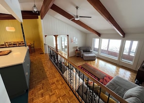 A spacious living room with a high ceiling, wooden beams, a dining area with chairs, a sofa, and a large window providing natural light.
