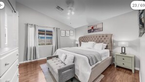 Een slaapkamer met een groot bed, een nachtkastje, een dressoir, een raam met gordijnen en een plafondventilator.