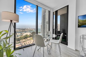 A modern room with a glass table, four chairs, a floor lamp, and a large window offering a city view. - Beautiful 2Bed Bay Views (Miami)