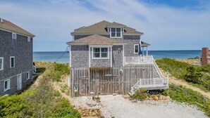 A two-story beach house with a wooden deck and a small porch, located near the ocean.