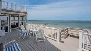 A wooden deck with white railings, a covered seating area with chairs and tables, and a view of the beach and ocean.