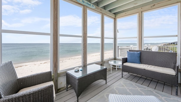 A covered outdoor seating area with wicker furniture, a glass table, and a view of the beach.