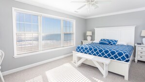 A bedroom with a large window, a bed with a blue patterned bedspread, a white ceiling fan, and a white nightstand with a lamp.