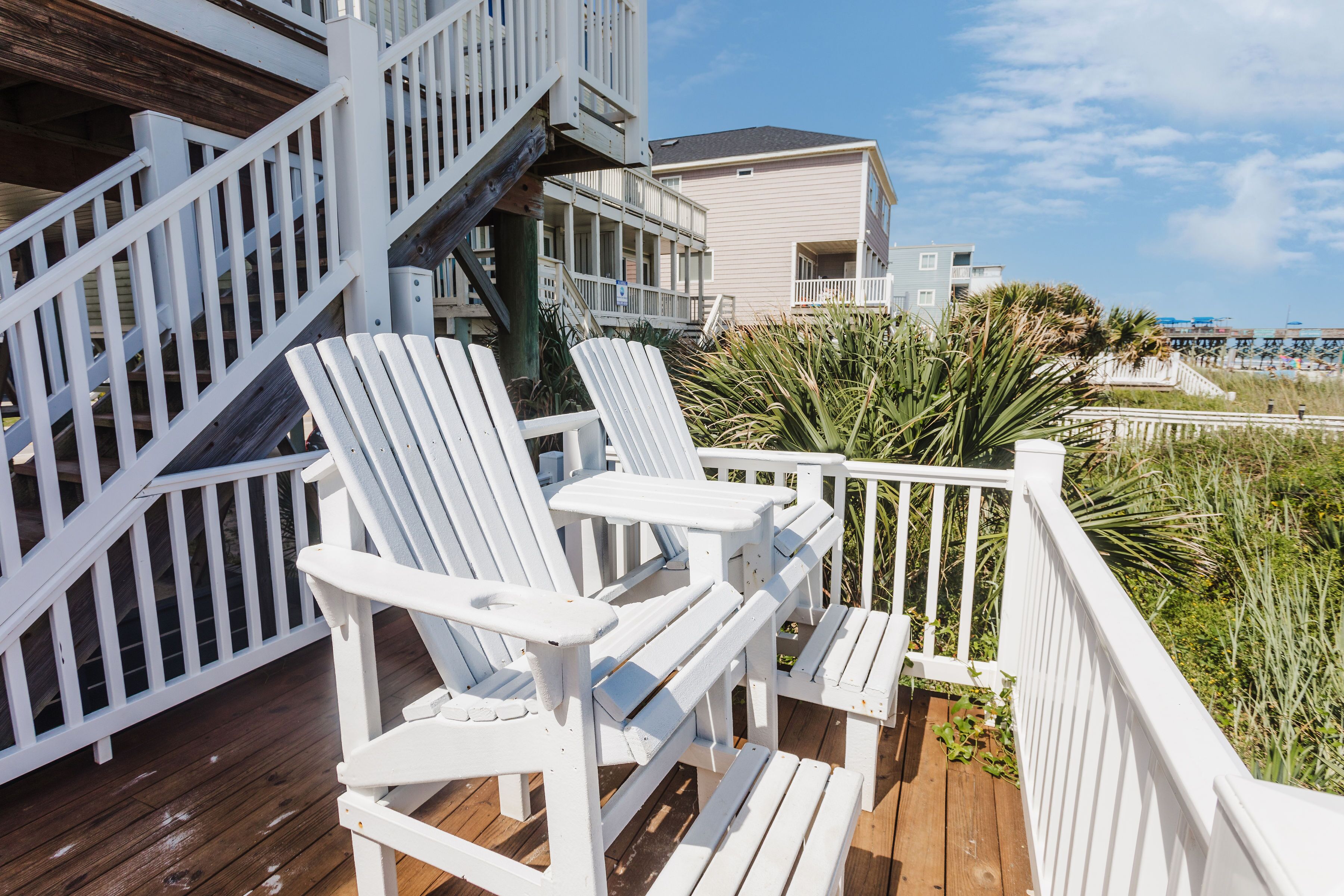 A wooden deck with white Adirondack chairs, a staircase, and a building with a balcony in the background.