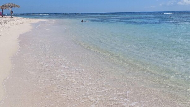Une plage avec une eau claire et peu profonde, quelques personnes au loin et un parasol en chaume sur la gauche.