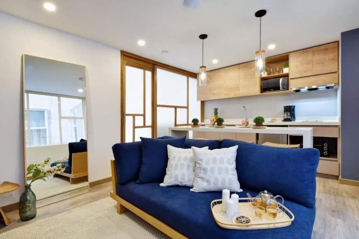 Un salon moderne avec un canapé bleu, une table basse sur laquelle sont posées des boissons, et une cuisine en arrière-plan.