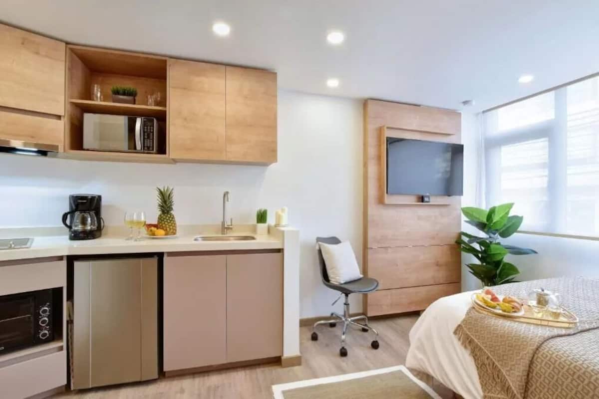 Une chambre d’hôtel compacte comprenant un coin cuisine, un lit avec un plateau de petit-déjeuner, un téléviseur fixé au mur et une plante en pot.