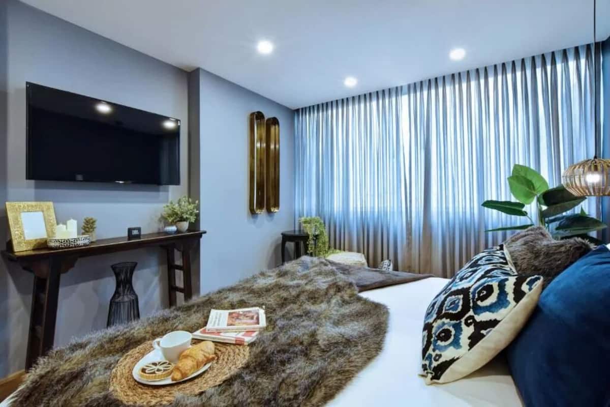 Une chambre moderne dotée d’un téléviseur à écran plat, d’une table de chevet avec une lampe, d’une grande fenêtre avec des rideaux et d’un lit confortable agrémenté d’un tapis moelleux.